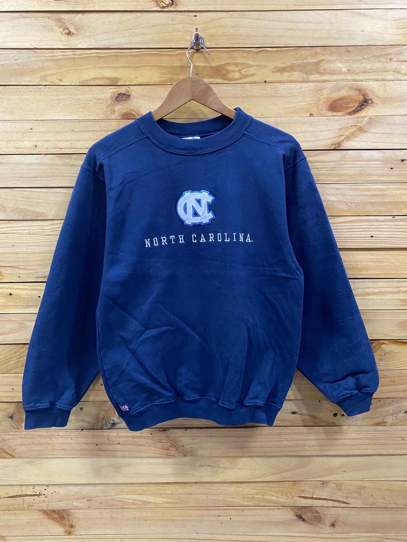 May include: Navy blue sweatshirt with the words "NORTH CAROLINA" in white lettering. The sweatshirt features a light blue and white logo on the chest. The sweatshirt is hanging on a wooden hanger.