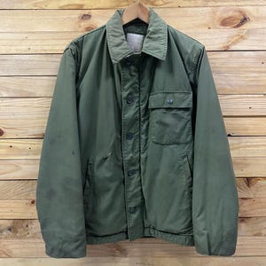 May include: Olive green military-style jacket with a button-up front, a collared neckline, and a single chest pocket. The jacket is hanging on a wooden hanger against a wooden plank background. The jacket has a worn appearance.