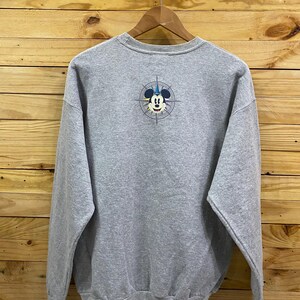 May include: A gray sweatshirt with a Mickey Mouse graphic on the back. The graphic features Mickey Mouse wearing a blue hat and a compass with a yellow, blue, and red design.