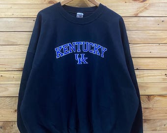 May include: A navy blue crewneck sweatshirt featuring the word KENTUCKY embroidered in blue with a white outline, positioned above a blue and white UK logo. The garment has long sleeves, a ribbed collar, and ribbed cuffs on a wooden hanger.