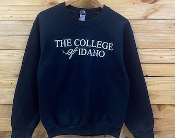 May include: A black long-sleeve crewneck sweatshirt featuring the white text THE COLLEGE of IDAHO printed across the chest. The garment has a ribbed collar, cuffs, and hem, and is displayed on a wooden hanger against a horizontal wood plank wall.