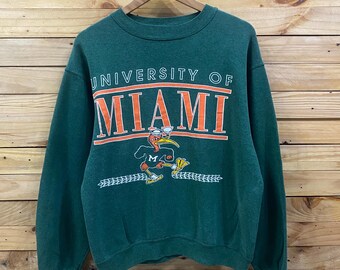 May include: A dark green sweatshirt with the words "UNIVERSITY OF MIAMI" in orange and white lettering. Below the text is a cartoon duck mascot in orange and white. The sweatshirt is hanging on a wooden hanger.
