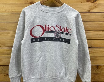 May include: A gray sweatshirt with the words "Ohio State University" printed on the front in red, black, and white. The sweatshirt is hanging on a wooden hanger against a wooden background.