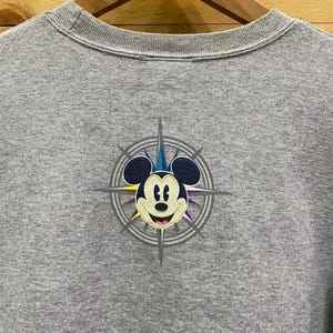 May include: Gray t-shirt with a graphic of Mickey Mouse in a compass design. The compass has a blue, yellow, and purple color scheme.