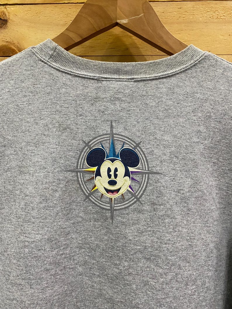 May include: Gray t-shirt with a graphic of Mickey Mouse in a compass design. The compass has a blue, yellow, and purple color scheme.