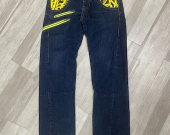 May include: Dark blue denim jeans featuring bright yellow pockets with a black graffiti-style print. The pants include two diagonal zippered pockets on the right thigh, a button fly, and a multicolored plaid fabric lining visible at the waistband.