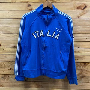 Könnte beinhalten: Blaue Trainingsjacke mit Reißverschluss, weißen Streifen an den Ärmeln und dem Wort "ITALIA" in weißen Blockbuchstaben auf der Brust. Drei weiße Sterne über dem "A".