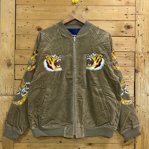 Pu&ograve; includere: Una giacca bomber in velluto a coste marrone con teste di tigre ricamate sul davanti e disegni di draghi sulle maniche.