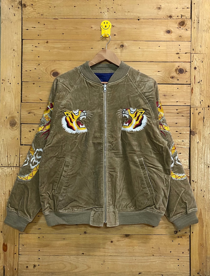 Pu&ograve; includere: Una giacca bomber in velluto a coste marrone con teste di tigre ricamate sul davanti e disegni di draghi sulle maniche.