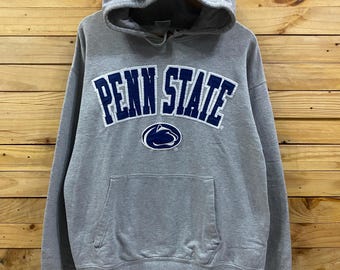 May include: A gray hooded sweatshirt with the words "PENN STATE" in navy blue arched across the chest. The sweatshirt has a front pocket and a Penn State logo below the text. The background is a wooden wall.