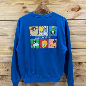 May include: A blue sweatshirt with a Looney Tunes design. The graphic features characters like Taz and Bugs Bunny. The words "LOONEY TUNES" are printed below the cartoon characters. The sweatshirt is displayed on a wooden hanger.
