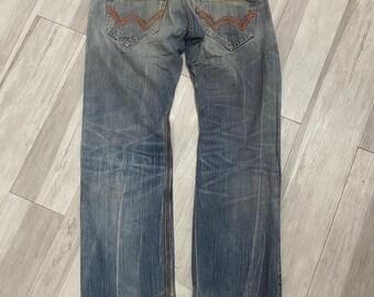 May include: Light blue denim jeans featuring a faded wash, a brown leather patch on the waistband, and two back pockets with orange zipper accents and decorative orange stitching. The hem of the pant legs shows signs of wear and fraying.