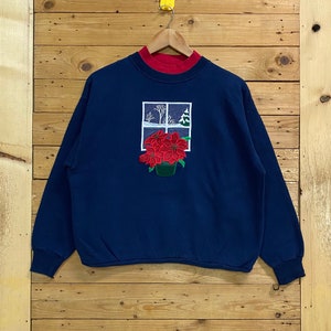 May include: A navy blue sweatshirt with a red ribbed collar and a winter scene embroidered on the front. The scene features a window with a snowy landscape and a potted poinsettia.