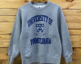 May include: A gray crewneck sweatshirt with the words "UNIVERSITY OF PENNSYLVANIA" printed in navy blue. The sweatshirt has a classic design and a ribbed collar, cuffs, and hem. The university's emblem is also printed on the front.