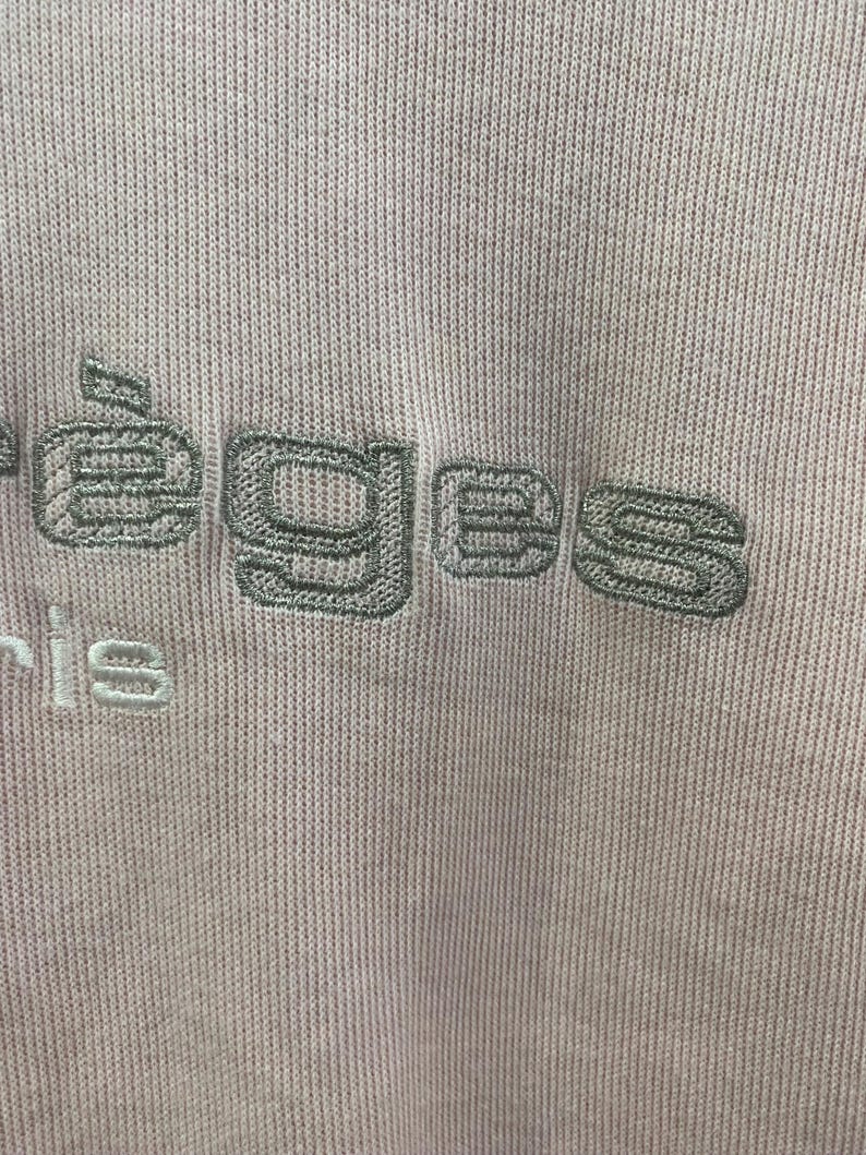 May include: Close-up of a light purple knit fabric with embroidered text. The text, in a muted green, reads "&eacute;eges" above "ris". The fabric has a ribbed texture, and the embroidery adds a subtle contrast in color and texture.