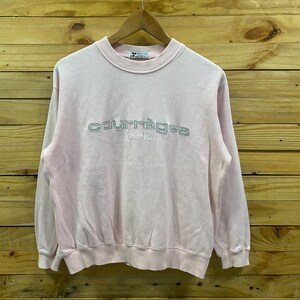 May include: A light pink Courr&egrave;ges Paris sweatshirt. The crew neck sweatshirt has the brand name embroidered in silver thread across the chest. The long-sleeved top is hanging on a wooden hanger against a wooden background.