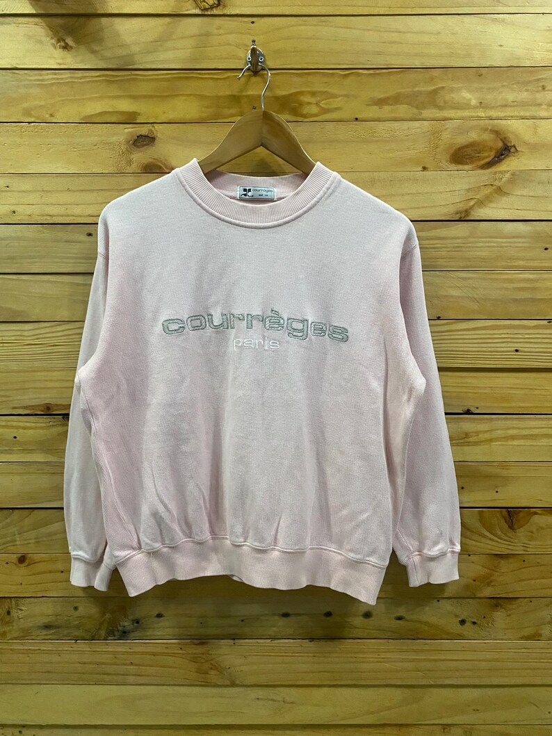 May include: A light pink Courr&egrave;ges Paris sweatshirt. The crew neck sweatshirt has the brand name embroidered in silver thread across the chest. The long-sleeved top is hanging on a wooden hanger against a wooden background.