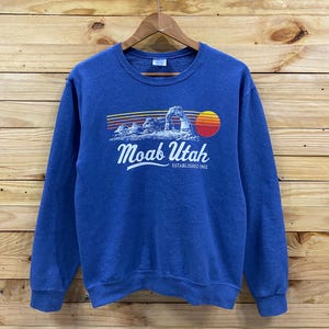 May include: Blue Moab Utah sweatshirt with a graphic of a desert landscape, a setting sun, and the text "Moab Utah ESTABLISHED 1902". The sweatshirt is hanging on a wooden hanger.