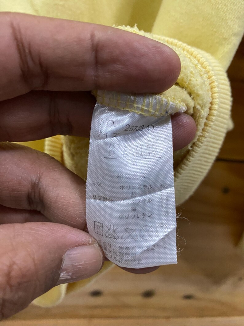 K&ouml;nnte beinhalten: Ein wei&szlig;es Etikett mit schwarzem Text, das die Gr&ouml;&szlig;e und die Stoffzusammensetzung eines gelben Sweatshirts angibt. Das Etikett lautet "NO. 25703-40", "バスト 79-87", "身長 154-162", "M", "ポリエステル 63%", "リサイクル 37%", "ポリウレタン 3%" und "洗濯表示".