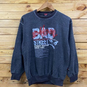 May include: Dark gray sweatshirt with the text "BAD STREET" in red and white, along with "STREET MASTER SINCE 1982". The sweatshirt has a crew neck and ribbed cuffs and hem. The garment is hanging on a wooden hanger.