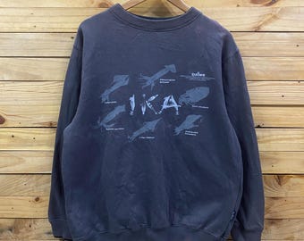 May include: A charcoal gray crewneck sweatshirt featuring a graphic print of various squid species and the text IKA in large letters. The design includes the Daiwa brand logo and small anchor icons, with ribbed cuffs and a ribbed hemline.