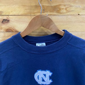 May include: Navy blue sweatshirt with the words "NORTH CAROLINA" embroidered in white. The sweatshirt features a light blue and white logo of the letters "NC". The sweatshirt is hanging on a wooden hanger.
