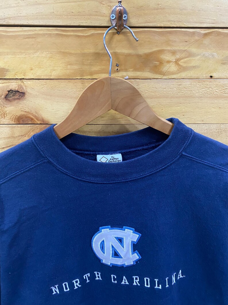 May include: Navy blue sweatshirt with the words "NORTH CAROLINA" embroidered in white. The sweatshirt features a light blue and white logo of the letters "NC". The sweatshirt is hanging on a wooden hanger.