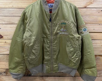 May include: Olive green bomber jacket with a ribbed collar, cuffs, and hem. The jacket features embroidered text and patches on the chest and sleeve. The jacket is hanging on a wooden hanger against a wooden background.