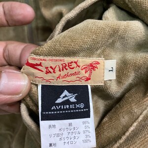 Pu&ograve; includere: Una giacca di velluto a coste marrone con chiusura a zip e un'etichetta che recita "ORIGINAL DESIGNS AVIREX Authentic". L'etichetta include anche una piccola grafica di palma. La giacca ha un'etichetta di taglia che recita "L". L'etichetta all'interno della giacca indica la composizione del tessuto: "表地 綿 95% ポリウレタン 5% リブ部分 アクリル 97% ポリウレタン 3% 裏地 ナイロン 100%"