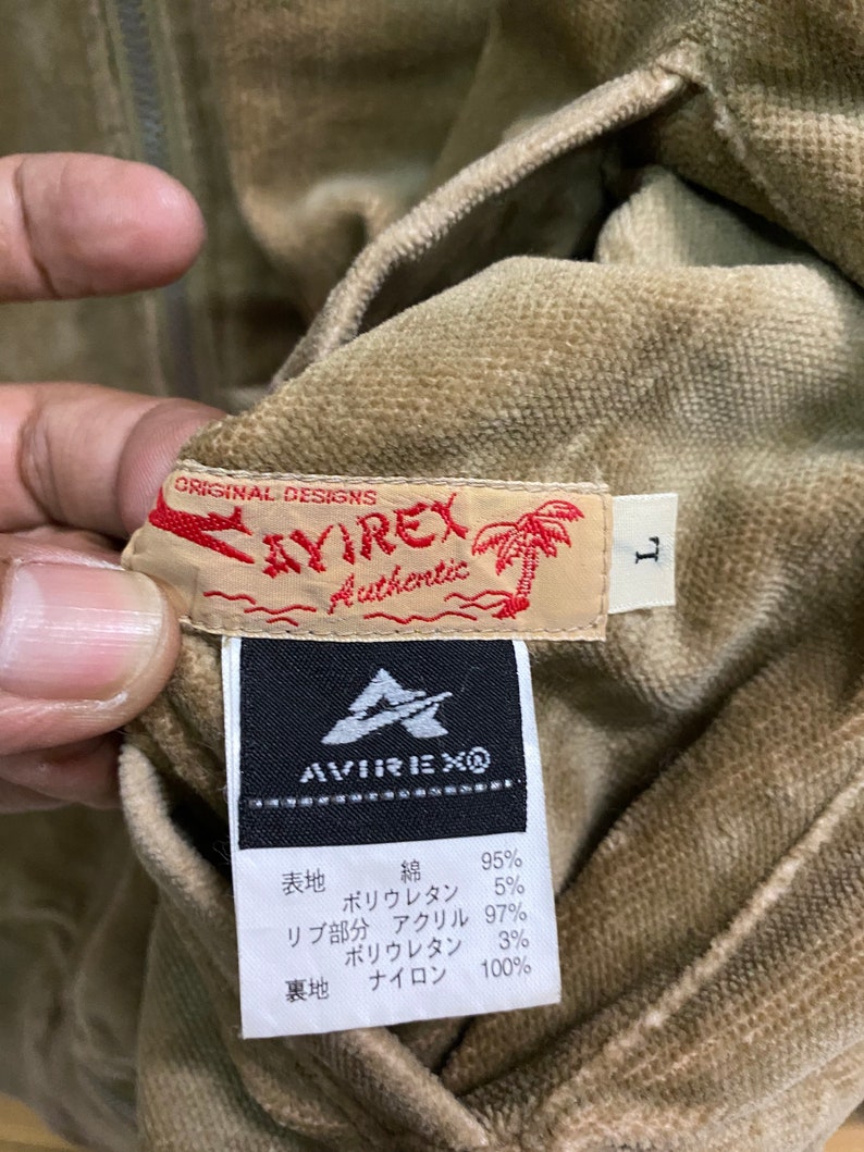 Pu&ograve; includere: Una giacca di velluto a coste marrone con chiusura a zip e un'etichetta che recita "ORIGINAL DESIGNS AVIREX Authentic". L'etichetta include anche una piccola grafica di palma. La giacca ha un'etichetta di taglia che recita "L". L'etichetta all'interno della giacca indica la composizione del tessuto: "表地 綿 95% ポリウレタン 5% リブ部分 アクリル 97% ポリウレタン 3% 裏地 ナイロン 100%"