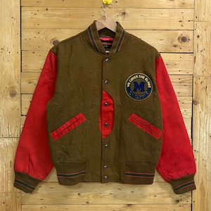 Könnte beinhalten: Eine braune und rote Collegejacke mit grünem und weißem Streifenbesatz. Die Jacke hat einen großen Aufnäher auf der linken Seite, auf dem "We Cover The Globe" steht.