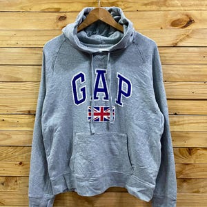 Puede incluir: Una sudadera con capucha GAP gris con capucha con cordón y bolsillo tipo canguro. La palabra "GAP" se muestra en letras azules con un contorno blanco, y una pequeña bandera de la Union Jack debajo.