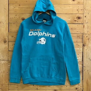 Puede incluir: Una sudadera con capucha azul turquesa con las palabras "Miami Dolphins" en letras naranja y blancas y un logotipo de casco de delfín.