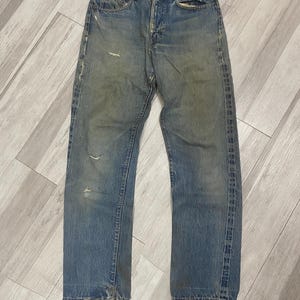 May include: Faded blue denim jeans with a classic five-pocket design. The jeans show signs of wear, including distressed areas and frayed edges. The straight-leg cut and button fly are visible.