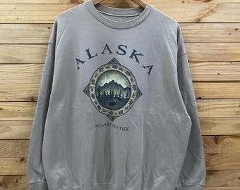 May include: A tan sweatshirt with the word "ALASKA" arched across the top. Below the word is a circular graphic with a mountain scene and the text "THE LAST FRONTIER". The sweatshirt is hanging on a wooden hanger.
