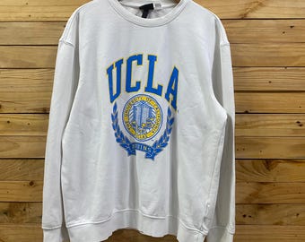 May include: White UCLA sweatshirt with the university's logo in blue and gold. The logo features the letters "UCLA" above a circular emblem with the words "University of California" and "Bruins". The sweatshirt is hanging on a wooden hanger.