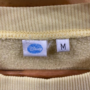 K&ouml;nnte beinhalten: Ein gelber Pullover mit einem blauen Disney-Logo und einem Gr&ouml;&szlig;enetikett M.