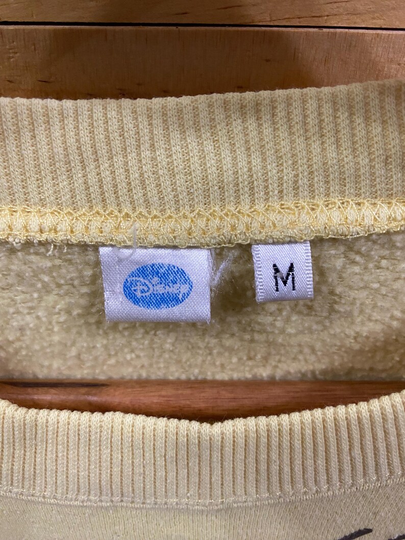 K&ouml;nnte beinhalten: Ein gelber Pullover mit einem blauen Disney-Logo und einem Gr&ouml;&szlig;enetikett M.