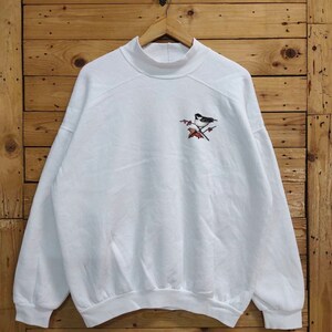 May include: A white long-sleeve sweatshirt featuring a mock neck collar and a small embroidered graphic on the upper left chest. The embroidery depicts a black and white bird perched on a brown branch with red berries and a single red leaf.