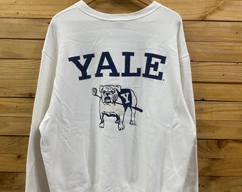 May include: White sweatshirt with the word "YALE" in navy blue block letters across the back. Below the word "YALE" is a navy blue graphic of a bulldog holding a stick. The sweatshirt is hanging on a wooden hanger.