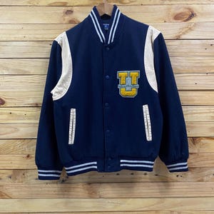 May include: A navy blue varsity jacket with cream-colored sleeves and accents. The jacket features a large yellow and gold letter "U" patch on the chest, along with white stripes on the collar, cuffs, and hem. The jacket is buttoned up and hanging on a wooden background.