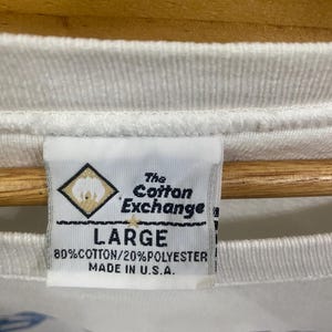 K&ouml;nnte beinhalten: Nahaufnahme eines wei&szlig;en T-Shirt-Etiketts. Auf dem Etikett steht "The Cotton Exchange" und "LARGE". Es besteht aus 80 % Baumwolle und 20 % Polyester, hergestellt in den USA. Das T-Shirt hat unten ein blaues Design.