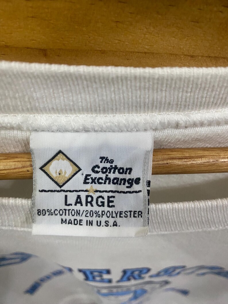K&ouml;nnte beinhalten: Nahaufnahme eines wei&szlig;en T-Shirt-Etiketts. Auf dem Etikett steht "The Cotton Exchange" und "LARGE". Es besteht aus 80 % Baumwolle und 20 % Polyester, hergestellt in den USA. Das T-Shirt hat unten ein blaues Design.