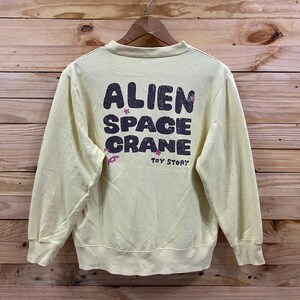 K&ouml;nnte beinhalten: Ein hellgelber Pullover mit einem grafischen Aufdruck auf der R&uuml;ckseite, der "Alien Space Crane" mit rosa Sternen und den Worten "Toy Story" darunter sagt.