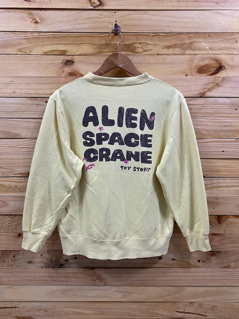 K&ouml;nnte beinhalten: Ein hellgelber Pullover mit einem grafischen Aufdruck auf der R&uuml;ckseite, der "Alien Space Crane" mit rosa Sternen und den Worten "Toy Story" darunter sagt.