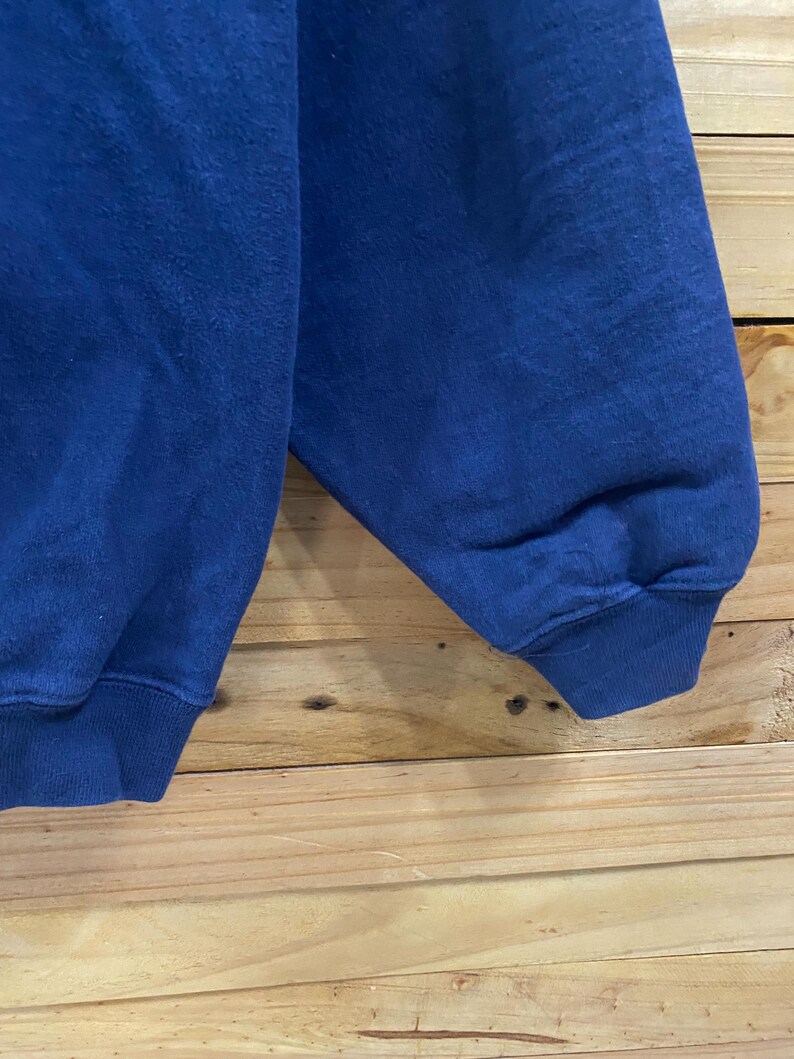 May include: Close-up of a blue sweatshirt sleeve. The sleeve is made of a soft, textured fabric and has a ribbed cuff. The background is a light brown wooden surface.