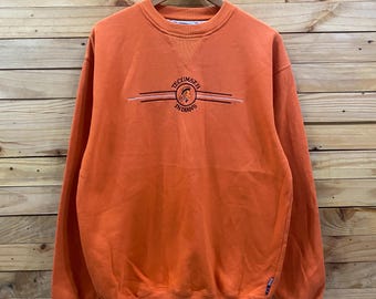 May include: Orange sweatshirt with the text "Tecumseh Indians" in a stylized design. The sweatshirt has a crew neck and long sleeves. The garment is hanging on a wooden hanger against a wooden background.