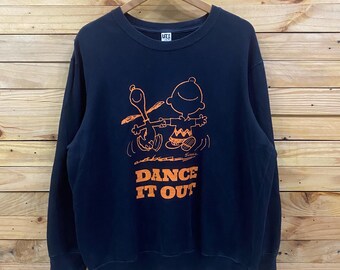May include: Black crew neck sweatshirt with an orange graphic of Charlie Brown and Snoopy dancing. The text "DANCE IT OUT" is below the characters. The sweatshirt is hanging on a wooden hanger against a wooden background.
