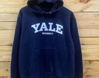 May include: Navy blue hooded sweatshirt with the word "YALE" in white block letters across the chest, with "UNIVERSITY" below. The sweatshirt has a front pocket and a drawstring hood. The background is a wooden wall.