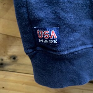 May include: Close-up of a navy blue garment with a ribbed cuff. A rectangular patch with the text "USA MADE" in red and white is sewn onto the garment. The background is a wooden surface.